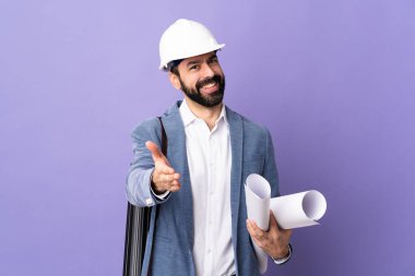 Miğferli genç bir mimar, izole edilmiş mor arka plan üzerinde planları tutuyor ve iyi bir anlaşma için el sıkışıyor.