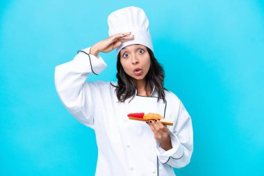 Genç İspanyol aşçı kadın mavi arka planda saşimi tutuyor. Yan tarafa bakarken sürpriz jest yapıyor.