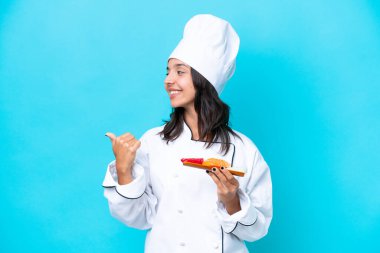 Mavi arka planda saşimi tutan genç İspanyol aşçı kadın bir ürünü sunmak için kenarı işaret ediyor.