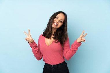 Mavi arka planda izole edilmiş genç melez kadın iki eliyle zafer işareti gösteriyor.