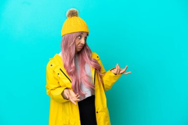 Pembe saçlı, yağmura dayanıklı bir ceket giyen genç bir kadın. Mavi arka planda yalıtılmış, sürpriz bir ifadeyle yan yana bakarken.