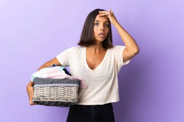 Mor arka planda izole edilmiş çamaşır sepetini tutan genç Latin kadın yan tarafa bakarken sürpriz jest yapıyor.