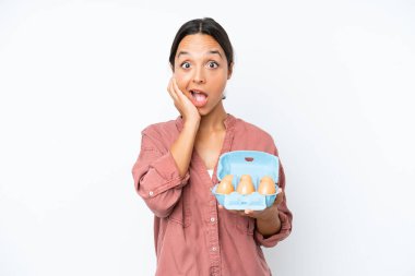 Beyaz arka planda yumurta tutan genç İspanyol kadın şaşırmış ve şok olmuş bir yüz ifadesiyle.