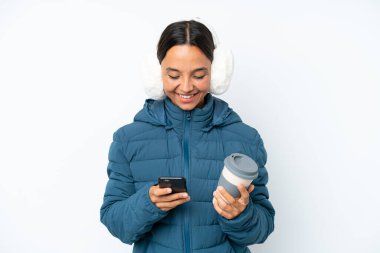Young hispanic woman wearing a winter earmuffs isolated on white background holding coffee to take away and a mobile