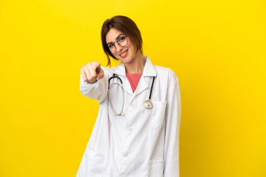 Doktor kadın sarı arka planda izole edilmiş. Parmağını kendine güvenen bir ifadeyle işaret ediyor.