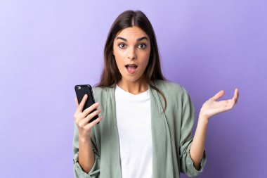 Mor arka planda izole edilmiş ve şok olmuş bir yüz ifadesi olan cep telefonu kullanan beyaz bir kadın.