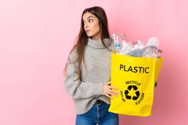 Pembe arka planda izole edilmiş bir şekilde geri dönüşüm için plastik şişelerle dolu bir çanta tutan beyaz bir kadın var.