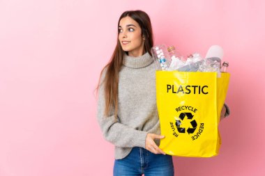 Pembe arka planda izole edilmiş bir şekilde geri dönüşüm için plastik şişelerle dolu bir çanta taşıyan beyaz bir kadın.