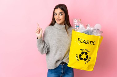 Pembe arka planda izole edilmiş plastik şişelerle dolu bir çanta taşıyan beyaz bir kadın bir ürünü sunmak için kenarı işaret ediyor.