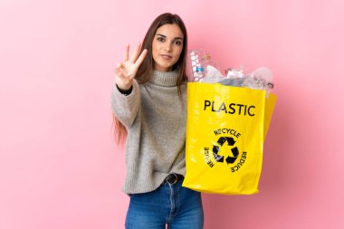 Pembe arka planda izole edilmiş plastik şişelerle dolu bir çanta taşıyan beyaz bir kadın mutlu ve parmaklarıyla üçe kadar sayıyor.