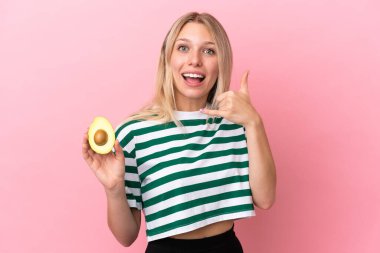 Avokado tutan beyaz bir kadın pembe arka planda izole edilmiş bir şekilde telefon jesti yapıyor. Bana geri dönüş işareti ver.