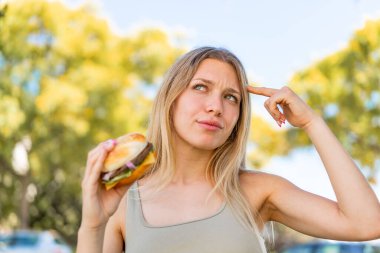 Genç sarışın kadın elinde hamburgerle kuşkuları ve kafası karışmış bir ifadeyle dışarıda bekliyor.