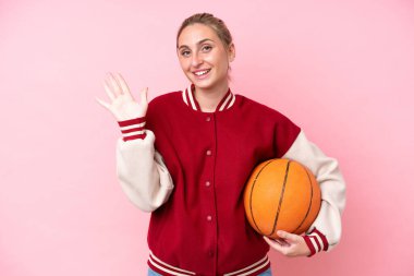 Genç basketbolcu beyaz kadın pembe arka planda izole edilmiş mutlu bir ifadeyle selam veriyor.