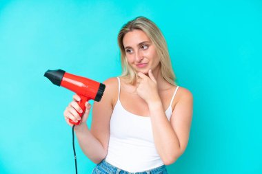 Elinde saç kurutma makinesi tutan beyaz bir kadın hüzünlü bir ifadeyle mavi arka planda izole edilmiş.