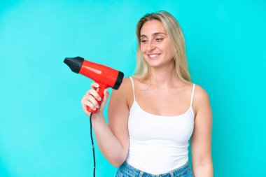 Kafkasyalı genç bir kadın elinde saç kurutma makinesiyle mavi arka planda mutlu bir ifadeyle izole edilmiş.