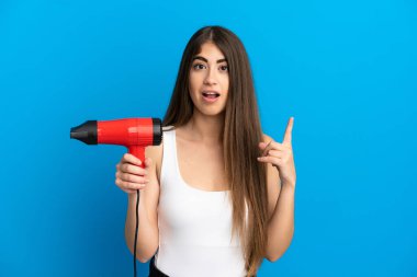 Kafkasyalı genç bir kadın elinde saç kurutma makinesiyle mavi arka planda izole edilmiş bir şekilde parmağını yukarı kaldıran bir fikir düşünüyor.