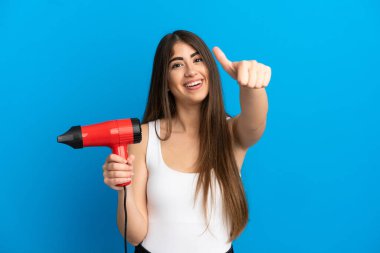 Young caucasian woman holding a hairdryer isolated on blue background with thumbs up because something good has happened
