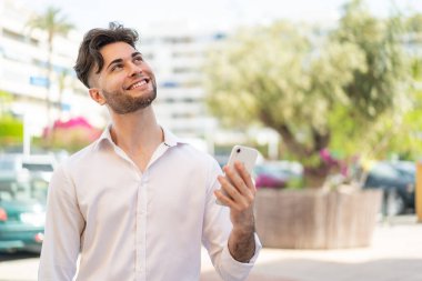 Dışarıda cep telefonu kullanan genç yakışıklı adam gülümserken yukarı bakıyor.