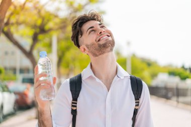 Elinde bir şişe su olan genç yakışıklı adam gülümserken yukarı bakıyor.