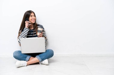 Yerde dizüstü bilgisayarı olan, beyaz arka planda izole edilmiş beyaz bir kadın elinde kahve ve bir cep telefonu ile oturuyor.