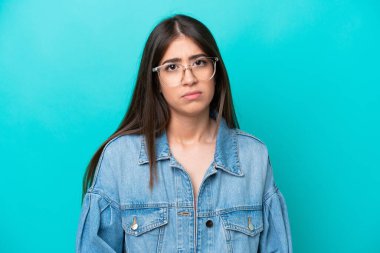 Mavi arka planda gözlük takan genç İspanyol kız şüpheci ve gergin, sorun yüzünden surat asıyor. negatif kişi.