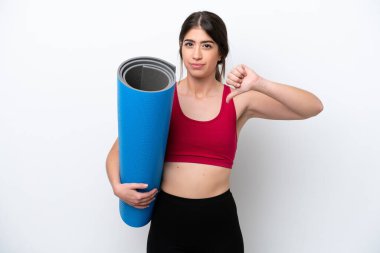 Genç sporcu kadın yoga derslerine giderken beyaz arka planda izole edilmiş bir minder tutuyor ve baş parmağını negatif ifadeyle aşağı gösteriyor.