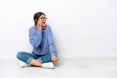 Beyaz arka planda izole bir şekilde yerde oturan genç beyaz kadın ağzı sonuna kadar açık bağırıyor.