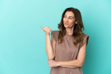 Mavi arka planda izole edilmiş orta yaşlı beyaz kadın bir ürünü sunmak için kenarı işaret ediyor.