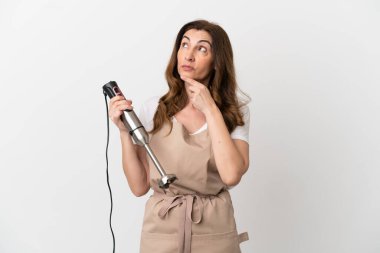 Beyaz arka planda izole edilmiş el blenderi kullanan orta yaşlı beyaz kadın gülümserken yukarı bakıyor.