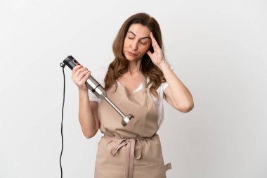 Orta yaşlı beyaz kadın el blenderi kullanıyor. Beyaz arka planda izole edilmiş, baş ağrısı var.