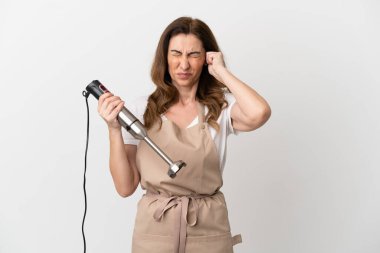 Beyaz arka planda izole edilmiş el blenderi kullanan orta yaşlı beyaz kadın hayal kırıklığına uğramış ve kulaklarını kapatmış.