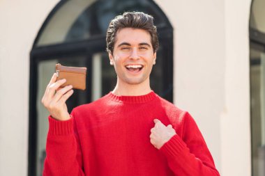 Dışarıda cüzdanını tutan yakışıklı bir genç. Yüzünde sürpriz bir ifade var.