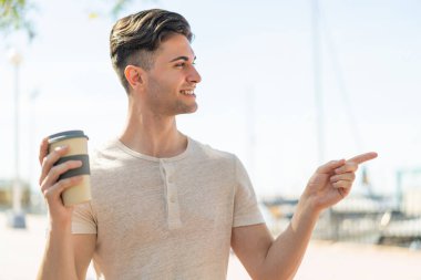 Genç ve yakışıklı bir adam açık havada kahvesini yudumlarken yan tarafı göstererek bir ürün sunuyor.