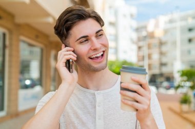 Genç yakışıklı adam cep telefonu kullanıyor ve elinde mutlu bir ifadeyle kahve tutuyor.