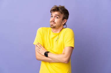 Genç yakışıklı sarışın adam mor arka planda izole edilmiş. Omuzlarını kaldırırken şüpheli hareketler yapıyor.