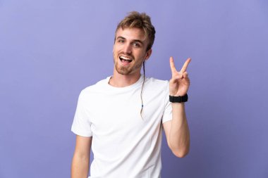 Mor arka planda izole edilmiş yakışıklı sarışın adam gülümsüyor ve zafer işareti gösteriyor.