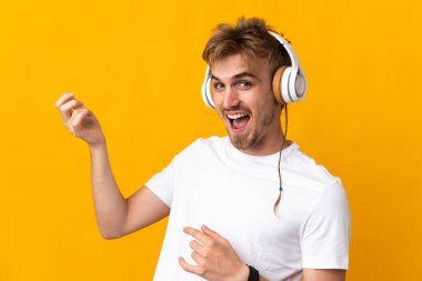 Sarışın genç adam sarı arka planda izole edilmiş müzik dinliyor ve gitar hareketi yapıyor.