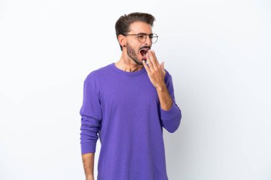 Beyaz arka planda izole edilmiş genç beyaz bir adam esniyor ve geniş bir ağzı eliyle kapatıyor.