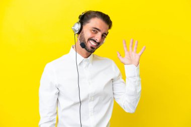 Tele pazarlamacı beyaz adam sarı arka planda izole edilmiş bir kulaklıkla çalışıyor mutlu bir ifadeyle selam veriyor.