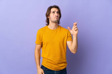 Mor arka planda izole edilmiş genç ve yakışıklı bir adam parmaklarını çapraz yapıp en iyisini diliyor.