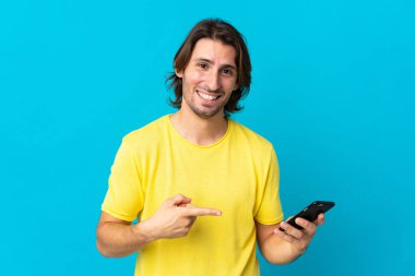Genç yakışıklı adam mavi arka planda izole edilmiş cep telefonu kullanıyor ve onu işaret ediyor.