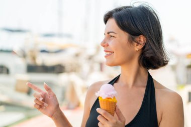 Açık havada kornet dondurması olan genç ve güzel Bulgar kadın bir ürün sunmak için kenarı işaret ediyor