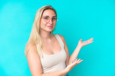 Mavi arka planda izole edilmiş genç beyaz kadın gözlüklü ve bir şeyler sunuyor.