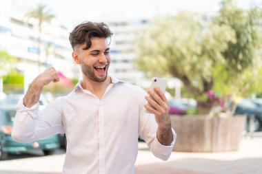 Dışarıda cep telefonu kullanan yakışıklı genç bir adam zaferi kutluyor.