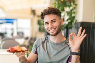 Genç yakışıklı adam açık havada sashimi tutuyor. Parmaklarıyla işaret veriyor.