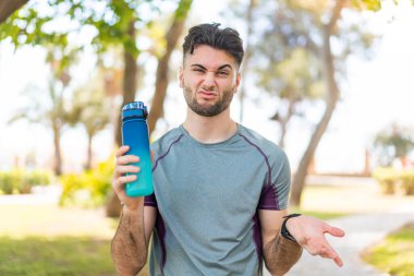 Genç yakışıklı sporcu dışarıda bir şişe su içerken omuzlarını kaldırırken şüpheyle hareket ediyor.
