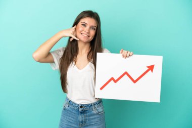 Mavi arka planda izole edilmiş genç beyaz bir kadın elinde büyüyen bir istatistik okuyla bir tabela tutuyor ve telefon jesti yapıyor.