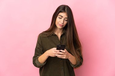 Pembe arka planda izole edilmiş genç beyaz kadın cep telefonuyla bir mesaj gönderiyor.