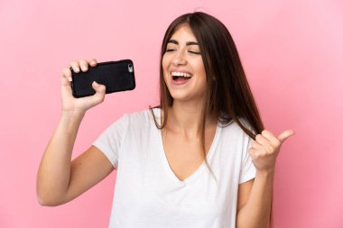 Pembe arka planda izole edilmiş genç beyaz kadın cep telefonu kullanıyor ve şarkı söylüyor.