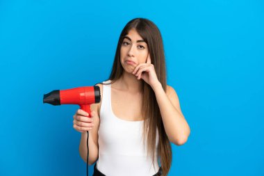 Kafkasyalı genç bir kadın elinde saç kurutma makinesiyle mavi arka planda izole edilmiş bir fikir düşünüyor.